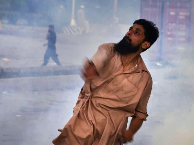 A Pakistani protester hurls back a tear gas canister fired by police, as another protester talks on a cell phone in the background