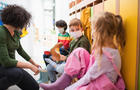 Pre school teacher helping children to put on shoes indoors in cloakroom at nursery, coronavirus concept. 